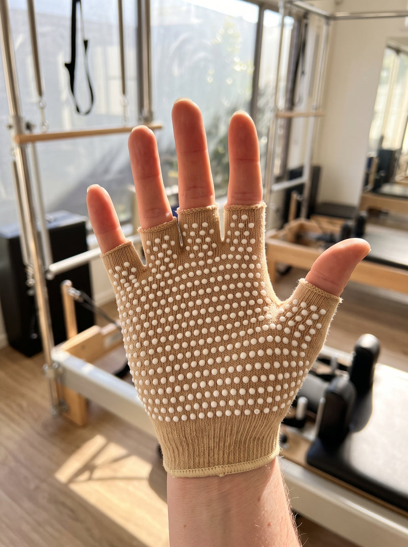 Open palm of an oat SoftGrip glove raised toward the camera at a reformer, dense raised silicone grip dots on palm and finger undersides, fingertips bare