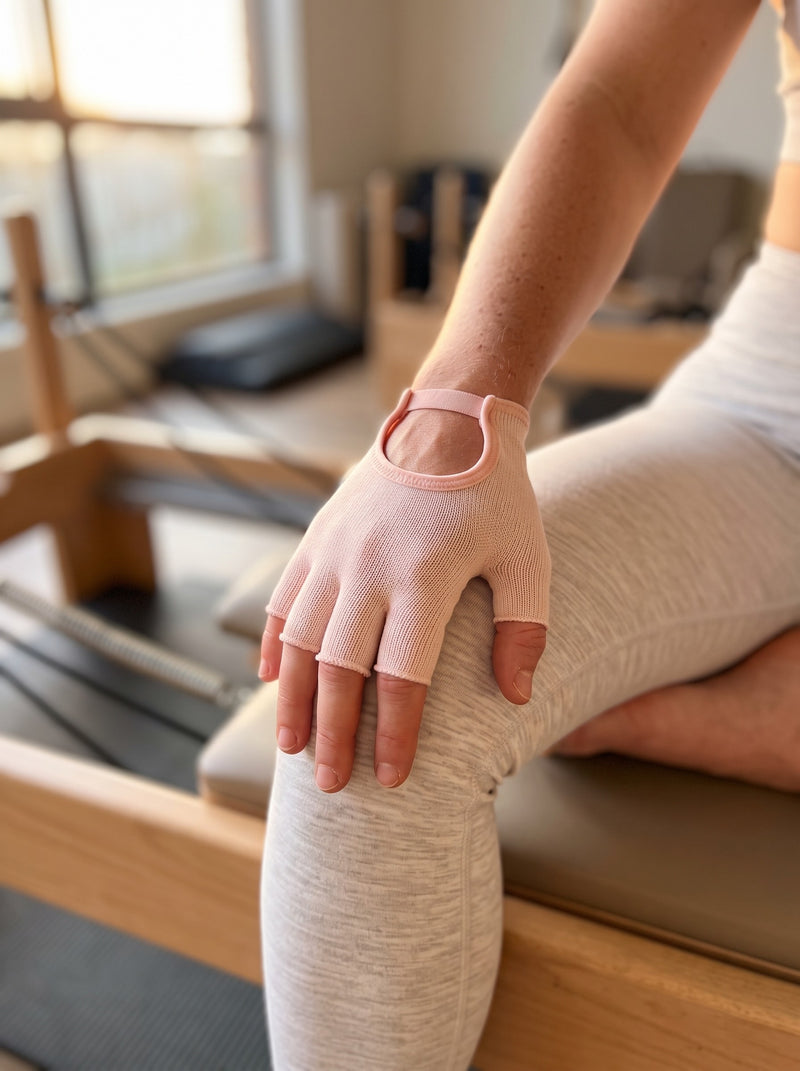 Back of a hand in a rosé SoftGrip glove resting on a knee at a Pilates reformer, open cut-out and wrist strap